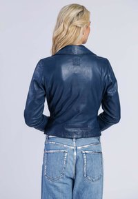 Giacca in pelle blu navy con una texture liscia, design aderente e un logo prominente impresso sulla schiena. Indossata con jeans azzurri.