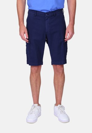 Homme portant un short cargo bleu marine, des baskets blanches et une chemise bleue, debout avec les mains relâchées le long du corps sur un fond uni.