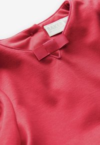 Top de tela roja con una textura suave y un pequeño lazo rectangular en el escote. La etiqueta dice "BAKER de Ted Baker."