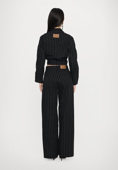 Juodas pinstripe kostiumas su prigludusia sakyte ir plačiomis kelnėmis. Yra diržo juosta ir tekstūruotas audinys. Šviesiai mėlynos juostelės pabrėžia dizainą.