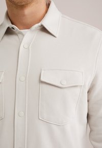 Primer plano de una persona que lleva una camisa abotonada color blanco roto con dos bolsillos de solapa en el pecho y un cuello puntiagudo.