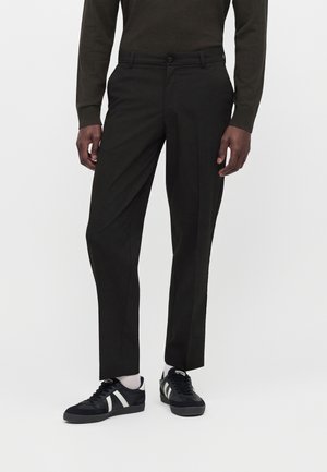 Mann, der schwarze maßgeschneiderte Hose, schwarzes Langarmhemd, weiße Socken und schwarze Sneaker mit weißen Streifen vor einem einfarbigen Hintergrund trägt.