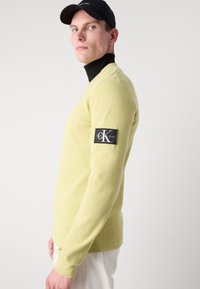 Lys gul langermet genser med strukturert strikk, med en svart lapp med hvit logo på venstre erme, båret over en svart høyhalset genser.