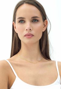 Guldkedja med en vågig design, med en texturerad yta. Bärs på en bar nacke och kompletterar en vit linne. Minimalistisk stil.