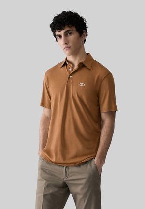 Giovane uomo con capelli ricci scuri che indossa una polo marrone a maniche corte e pantaloni beige, in piedi con le mani nelle tasche davanti a uno sfondo grigio.