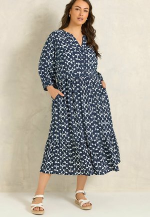 Woman wearing a navy blue and white patterned long-sleeve midi dress with pockets and white sandals, standing against a plain light background.