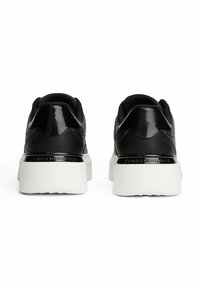 Un paio di sneakers nere con dettagli lucidi neri sul tallone e suole bianche spesse, viste da dietro su uno sfondo bianco.