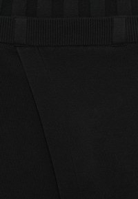 Melna ribota auduma tuvplāns, kurā redzama teksturēta adīšana, elastīga josta un trīsstūra formas detaļa, kas nodrošina strukturētu izskatu.