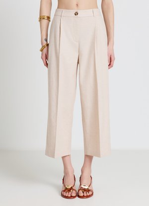 Femme portant un pantalon beige taille haute, court, large avec des plis, des sandales marron ouvertes à talons, des bracelets dorés et un petit tatouage au poignet.