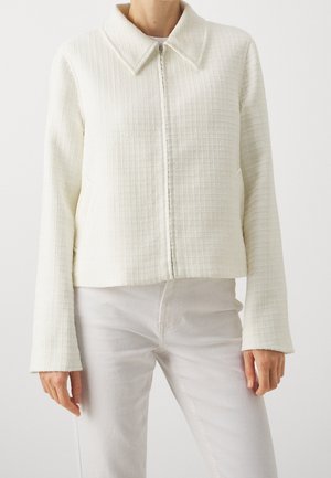 Femme portant une veste zippée écrue texturée avec un large col et des manches longues, associée à un pantalon de couleur claire.