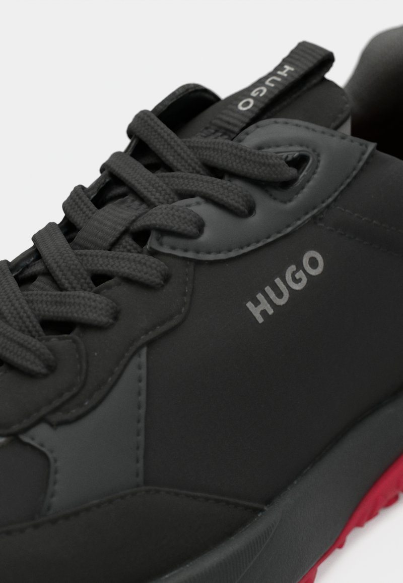 Schwarze Sportschuhe mit strukturiertem, synthetischem Obermaterial, auffälligem Logo, runden Schnürsenkeln und kontrastierender roter Sohle. Glatte Oberfläche mit minimalen Akzenten.
