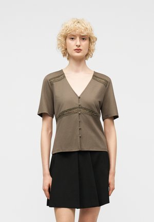 Vaaleat, lyhyet ja kiharat hiukset omaava nainen pukeutuu oliivinvihreään V-kaulaiseen lyhythihaiseen toppiin, jossa on napit, ja mustaan laskostettuun hameeseen yksinkertaista taustaa vasten.