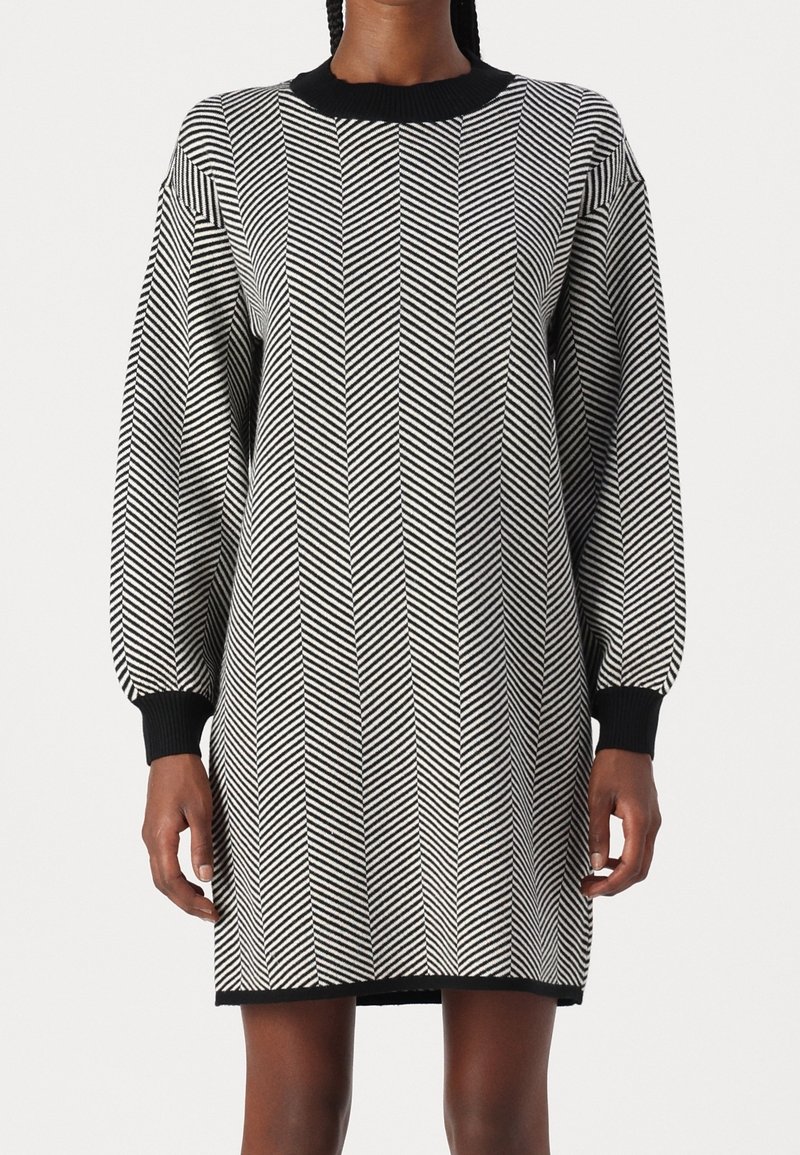 Robe en maille à manches longues avec motif chevron noir et blanc, col, poignets et ourlet noirs côtelés, portée par une personne debout.