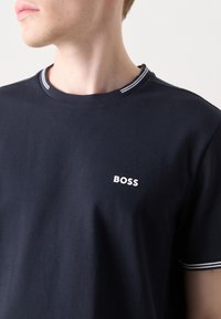 Tamsiai mėlynas kūno formos marškinėliai iš medvilnės. Ant krūtinės yra baltas "BOSS" logotipas ir juostelė ant apykaklės.
