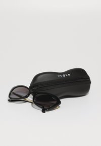 Óculos de sol pretos em forma de gato com lentes degradê, acompanhados de um estojo preto com fecho éclair que exibe o logótipo "Vogue" em branco.