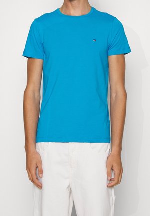 Mand iført en lyseblå T-shirt med korte ærmer og en lille logo på brystet samt hvide bukser, stående mod en hvid baggrund.