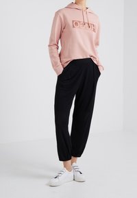 Sudadera con capucha de color rosa claro con un logo brillante, combinada con pantalones holgados negros y zapatillas blancas, sobre un fondo liso.