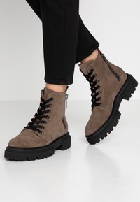 Bottes chelsea en suède marron avec lacets noirs et semelle noire robuste, dotées d'une fermeture éclair latérale et d'une finition texturée.