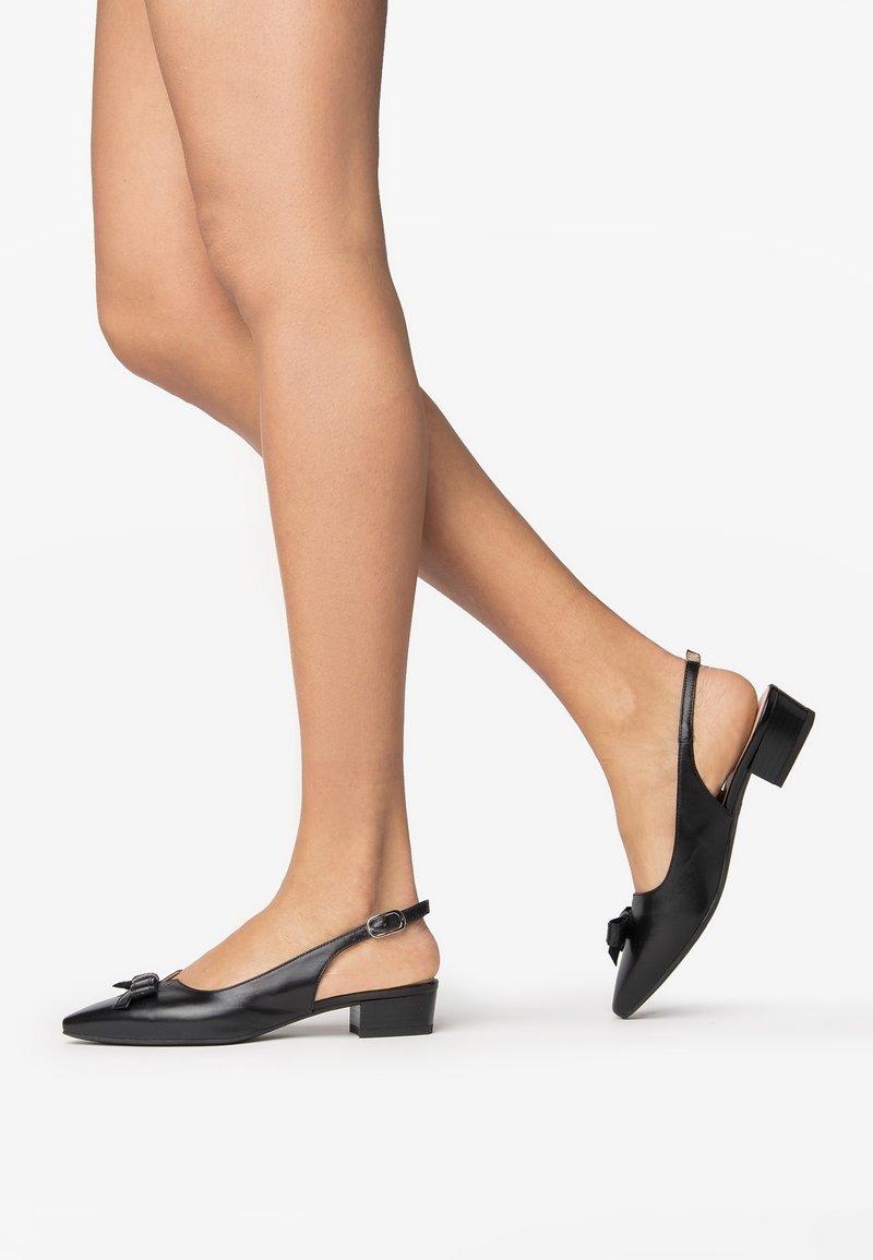 Jambes portant des chaussures slingback en cuir noir avec des bouts pointus, des petits talons carrés et un nœud à l'avant, sur un fond blanc uni.