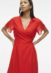 Rochie roșie înfășurată, cu mâneci scurte și evazate, având o textură în zigzag și un cordon la talie. Materialul este ușor și puțin transparent.
