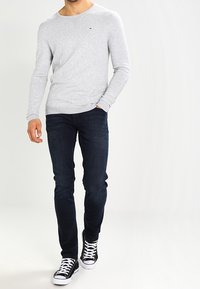 Homme portant une chemise à manches longues gris clair, un jean bleu foncé et des baskets noires et blanches, marchant avec une main dans la poche.