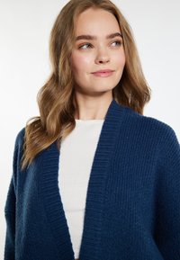 Mornarsko modra pletena jakna z rebrasto teksturo, odprta spredaj in z širokimi rokavi, nosi se preko kreme barvane srajce. Mehak, debel material.