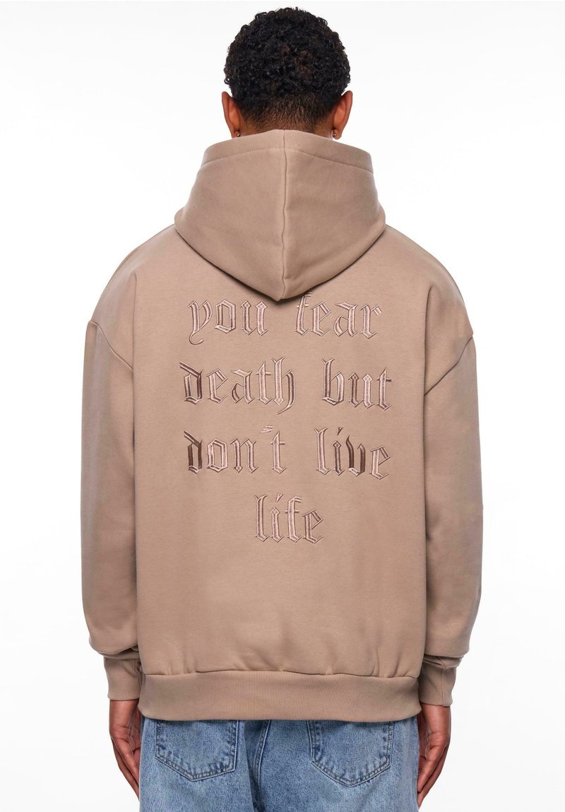 Sweat-shirt à capuche beige avec une broderie au dos en texte sombre : "tu crainds la mort mais tu ne vis pas la vie." Poche kangourou et poignets côtelés.