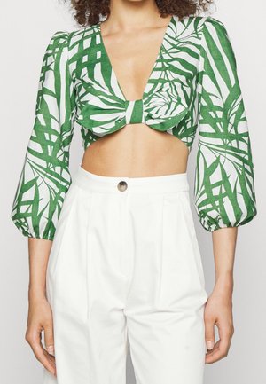 Blouse courte imprimé tropical vert et blanc avec manches trois-quarts bouffantes, associée à un pantalon blanc taille haute fermé par un bouton.