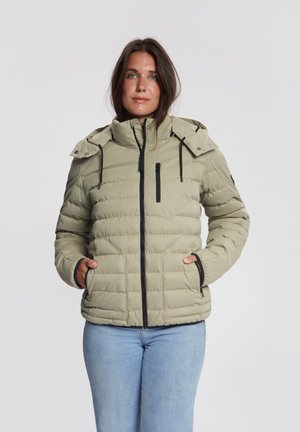 Vrouw met donker haar die een licht olijfgroene doorgestikte jas met capuchon en lichtblauwe spijkerbroek draagt, staat met handen in de jaszakken.