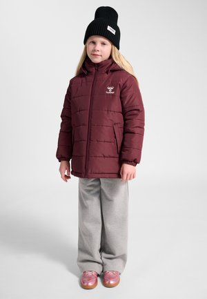 Barn iført en burgundy quiltet jakke, grå bukser, pink sko og sort strikhue, stående mod en ensfarvet lys baggrund.