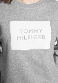 Gray sweatshirt with a white rectangular patch featuring the text "TOMMY HILFIGER" in embroidered gray letters. Soft texture.