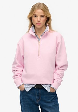 Femme portant un sweat-shirt à fermeture éclair quart de couleur rose clair sur une chemise rayée bleue et un jean bleu, debout les mains dans les poches devant un fond uni.