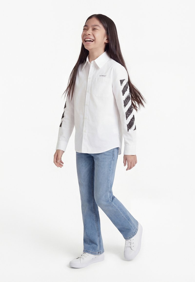 Κορίτσι με μακριά σκούρα μαλλιά που χαμογελά, περπατάει, φοράει άσπρο πουκάμισο με κουμπιά και μαύρες διαγώνιες ρίγες στα μανίκια, ανοιχτόχρωμο μπλε τζιν και λευκά αθλητικά παπούτσια.