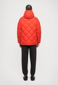 Red quilted puffer jacket with a high collar and hood, featuring elastic cuffs. Worn over dark trousers and black shoes, viewed from behind.