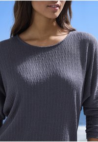 Frau, die ein gemustertes graues Langarmshirt trägt, steht im Freien vor einem klaren blauen Himmel.