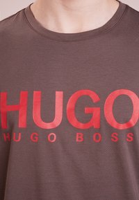 Camiseta de color marrón oscuro con el texto grande en rojo "HUGO" y el texto más pequeño "HUGO BOSS" debajo, cuello redondo y textura de tela suave.