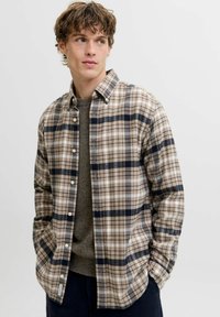 Młody mężczyzna z kręconymi włosami, ubrany w beżową, granatową i białą koszulę w kratę zapinaną na guziki, na szarym swetrze, z rękami w kieszeniach, patrzący w bok.