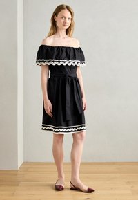 Čierne šaty bez ramienok s bielym lemovaním v tvare vlny, naskladaný živôtik, definovaný pás a opasok na páse. Modelka má na sebe bordové balerínky.