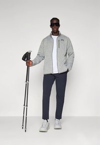 Harmaa fleece-takki vetoketjulla, valkoinen t-paita, tummat housut ja vaaleat tennarit. Malli pitää kädessään vaelluskeppiä ja käyttää aurinkolaseja. Yksinkertainen tausta.