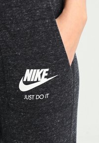 Tmavě šedé tepláky vyrobené z texturované látky, s bílým logem Nike a nápisem "JUST DO IT" potisknutým na oblasti přední kapsy.