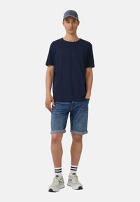T-paita tummansinistä, lyhythihainen, farkkushortsit, valkoiset sneakersit harmailla yksityiskohdilla, ja mustavalkoiset raidalliset sukat, valkoista taustaa vasten.