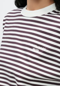 Κοντινό πλάνο ενός ατόμου που φο ράει πουκάμισο με λευκές και σκούρες μωβ ρίγες, με λευκή ριμπ λαιμόκοψη και κεντημένο λογότυπο "Carhartt WIP" στο στήθος.