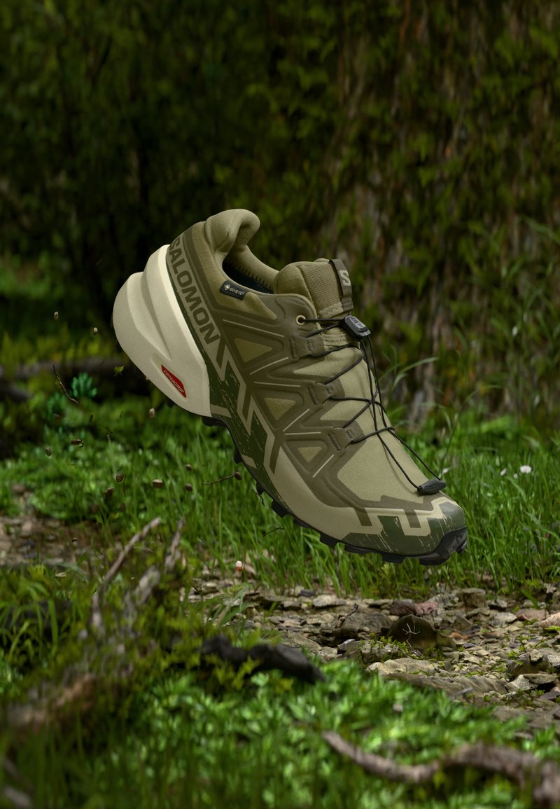 Ανοιχτό πράσινο παπούτσι Salomon για τρέξιμο σε μονοπάτια, αιωρούμενο πάνω από το δάπεδο ενός δάσους με γρασίδι, βράχους και κορμούς δέντρων στο φόντο.