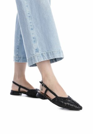 Zapatos de slingback trenzados en negro con punta afilada, tacón plano y correa ajustable en el tobillo, combinados con jeans de pierna ancha de color azul claro.