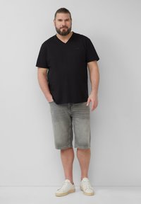 Czarny t-shirt z dekoltem w serek, szare szorty dżinsowe, białe sneakersy z beżowymi podeszwami. Model stoi swobodnie z rękami w kieszeniach na jasnym tle.