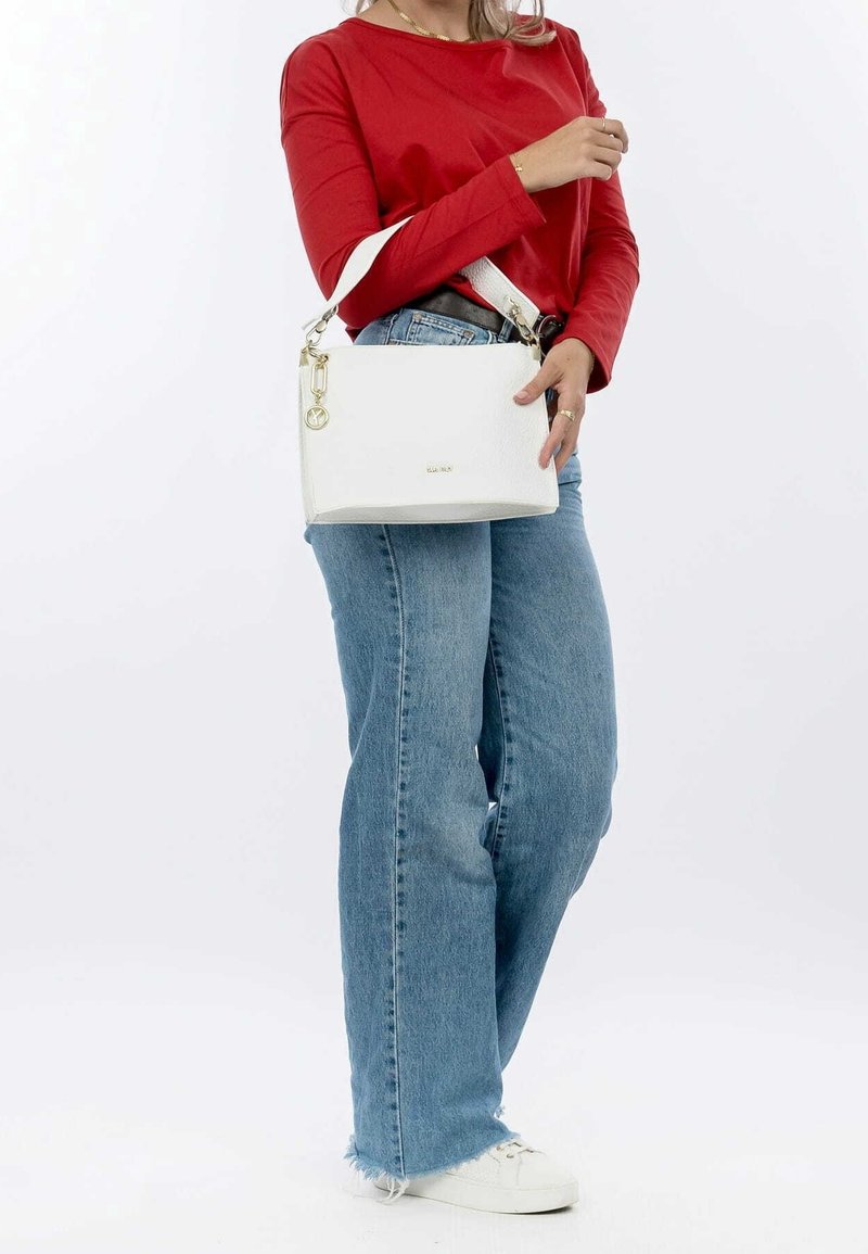 Femme portant un chemisier rouge à manches longues et un jean bleu, tenant un sac à main blanc avec des touches dorées et des baskets blanches sur un fond uni.