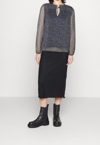 Blusa nera a maniche lunghe con sovrapposizione trasparente e testurizzata, dotata di un piccolo nodo frontale, abbinata a una gonna nera aderente e stivali neri alla caviglia chunky.