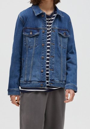 Personne portant une veste en jean bleu sur un chemisier rayé bleu marine et blanc, un pantalon gris et des colliers en argent superposés sur un fond uni.