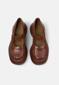 Chaussures synthétiques brunes avec une semelle en caoutchouc épaisse et une lanière de style Mary Jane. Présente des nervures texturées au niveau des orteils et une finition lisse.