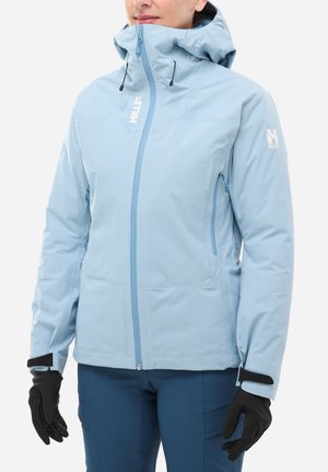 Femme portant une veste imperméable à capuche bleu clair avec fermeture éclair frontale, gants noirs et pantalon bleu foncé sur un fond blanc.